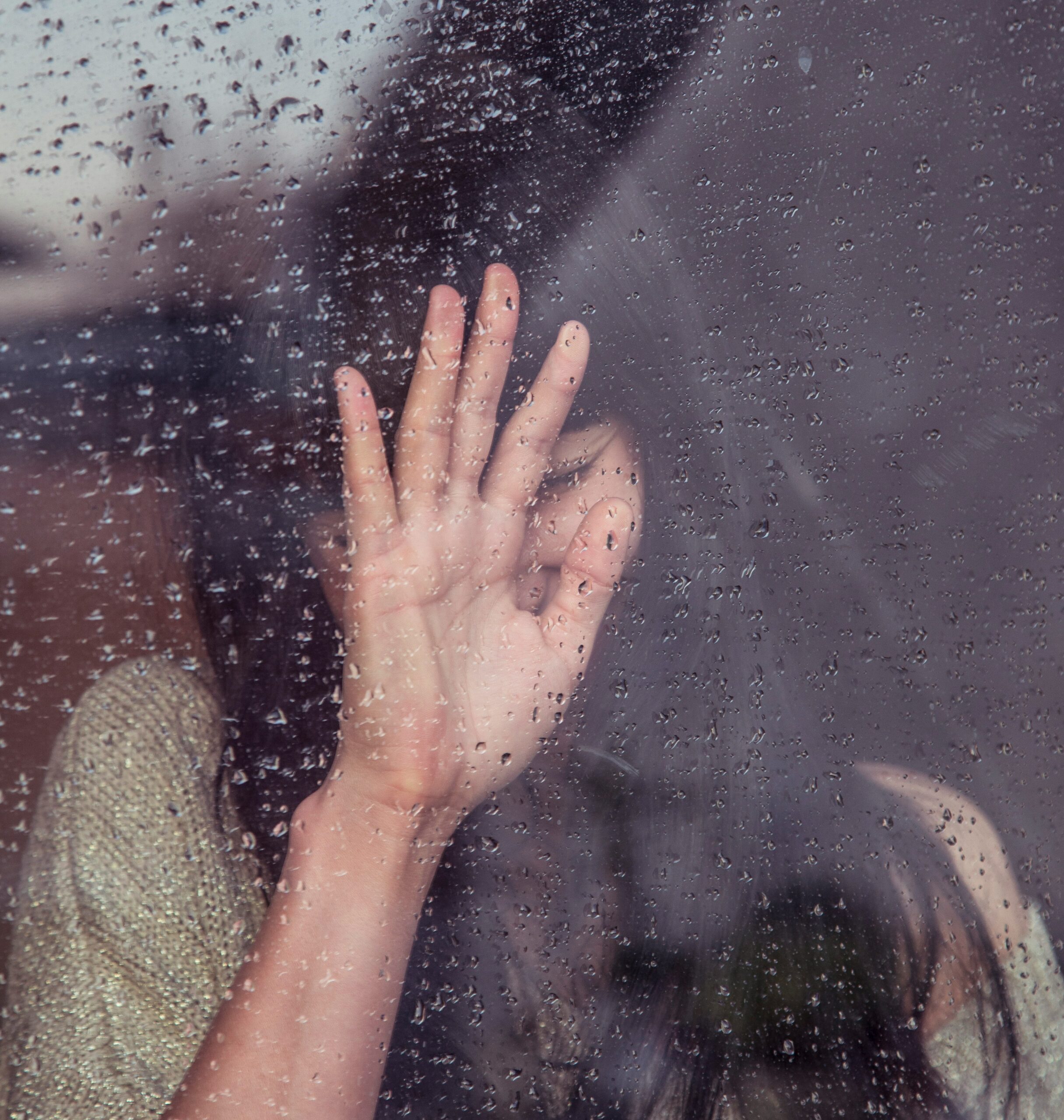 A lady experiencing greif behind a window with her hand up to the glass, rain covers the the whole image.